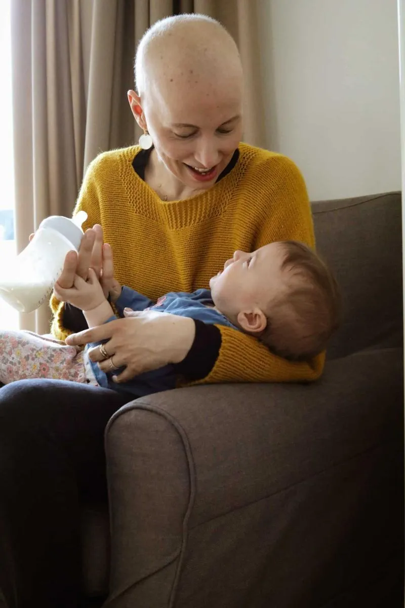Frau mit kahlem Kopf sitzt im Sessel und gibt einem Baby auf ihrem Schoß lächelnd das Fläschchen.