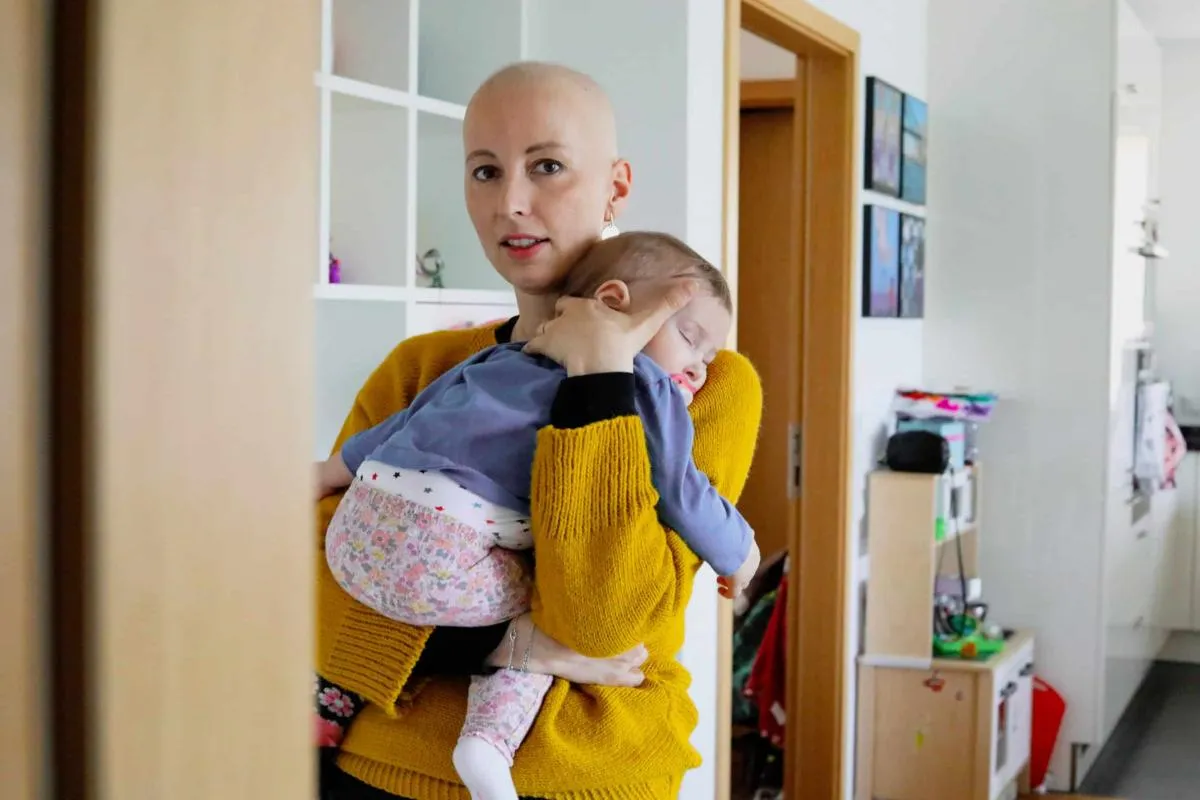 Frau mit kahlem Kopf trägt ein schlafendes Kleinkind auf dem Arm und blickt in die Kamera.