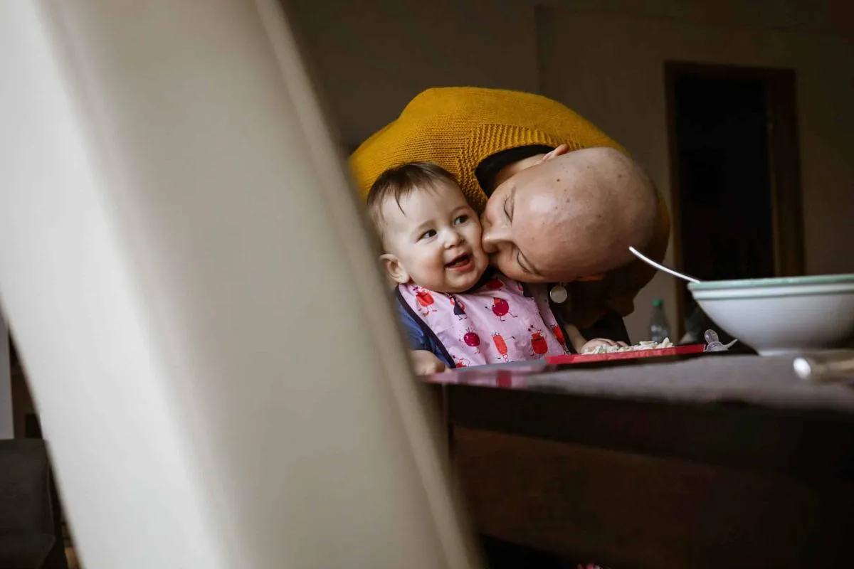 Frau mit kahlem Kopf küsst am Esstisch liebevoll die Wange eines lachenden Babys.