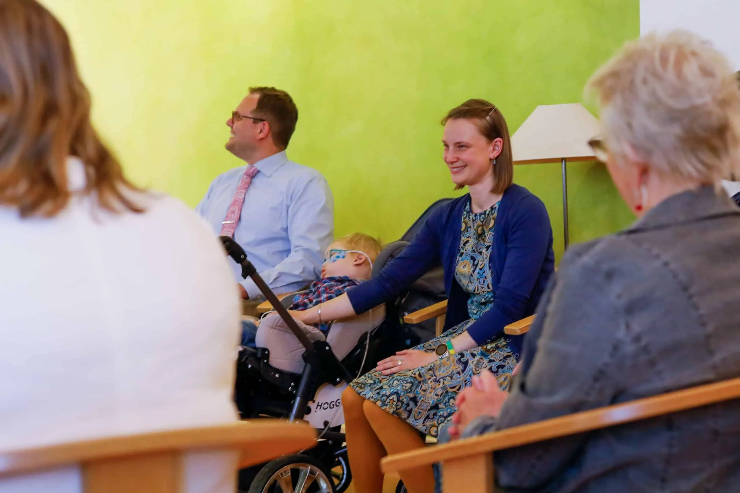 Die Eltern sitzen lächelnd neben ihrem Kind, das während der Tauffeier ruhig in seinem Buggy liegt.