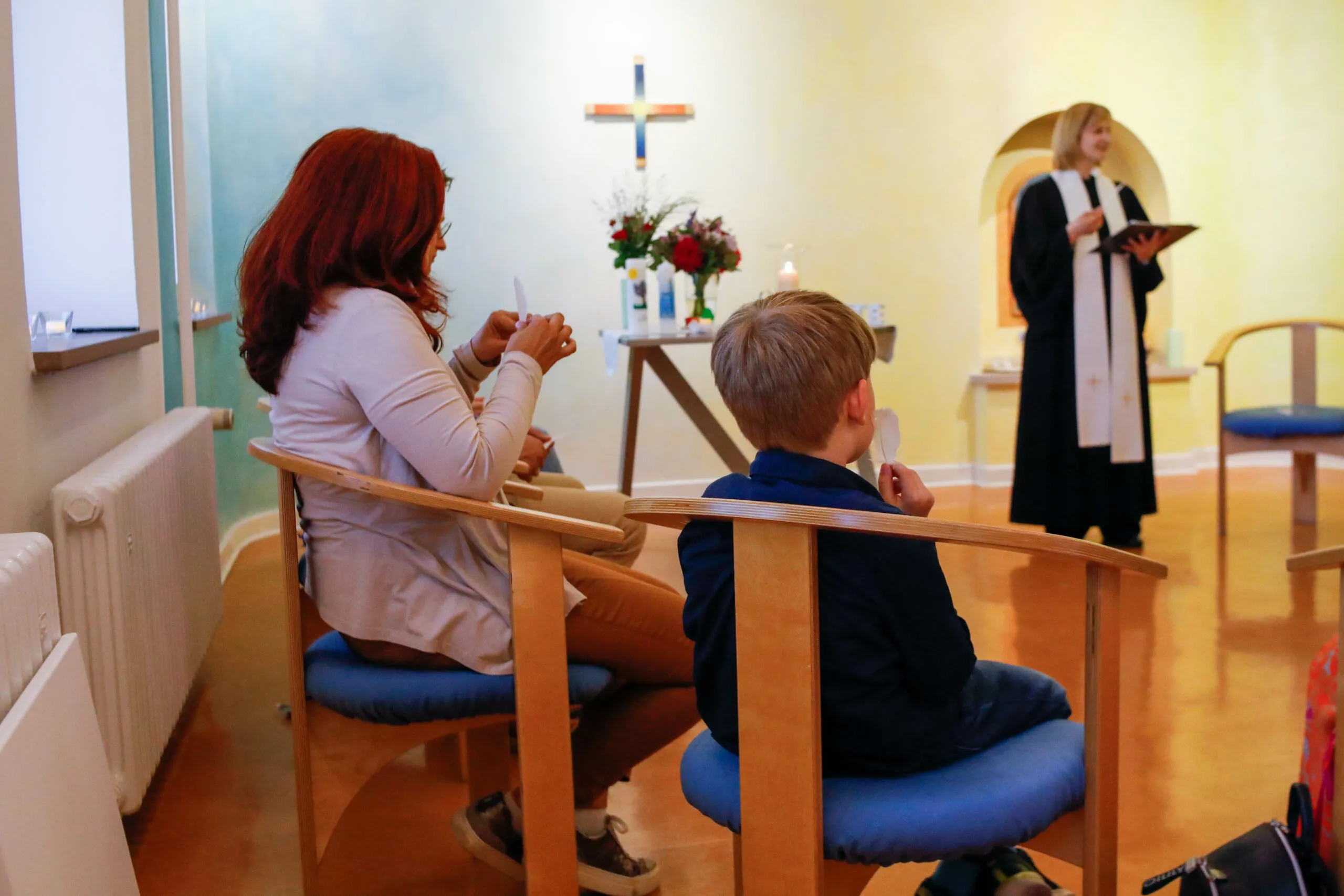 Die Familie sitzt in der Kapelle vor Kreuz, Blumen und Taufkerzen, während die Pfarrerin spricht.