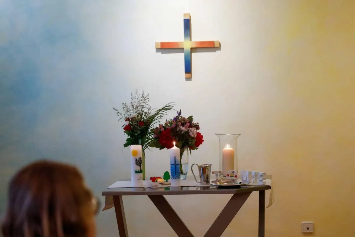 Kleiner Tisch mit Blumen und brennenden Kerzen unter einem bunten Holzkreuz an der Wand.