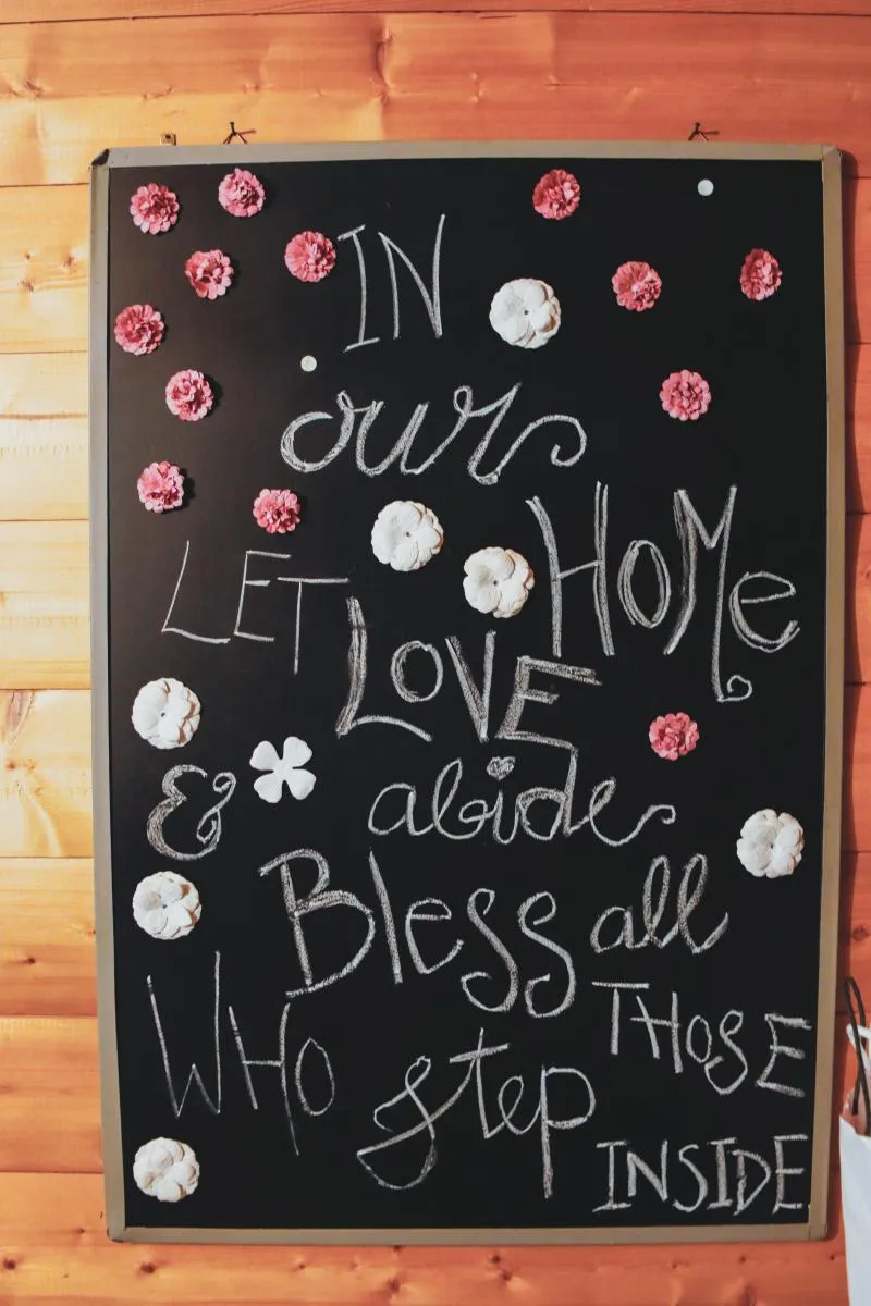 Kreidetafel mit Blütendeko und englischem Spruch: In our home let love abide, bless all those who step inside.