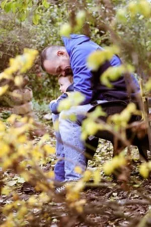 Vater umarmt sein Kind innig auf einer Bank im herbstlichen Garten, davor gelb blühende Zweige.