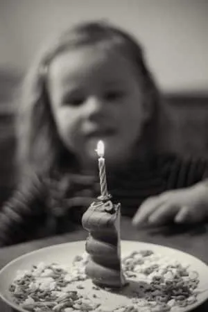 Schwarz-Weiß-Bild: Kleines Kind betrachtet eine brennende Kerze auf einem kleinen Kuchen mit bunten Streuseln.