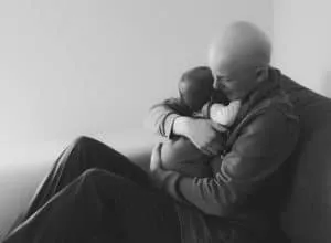 Schwarz-Weiß-Bild: Mann ohne Haare sitzt auf einem Sofa und hält ein Baby eng an seine Brust geschmiegt.