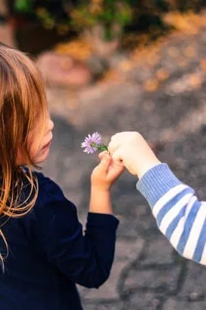 Eine Kinderhand reicht einem kleinen Mädchen draußen eine zarte lila Blume.