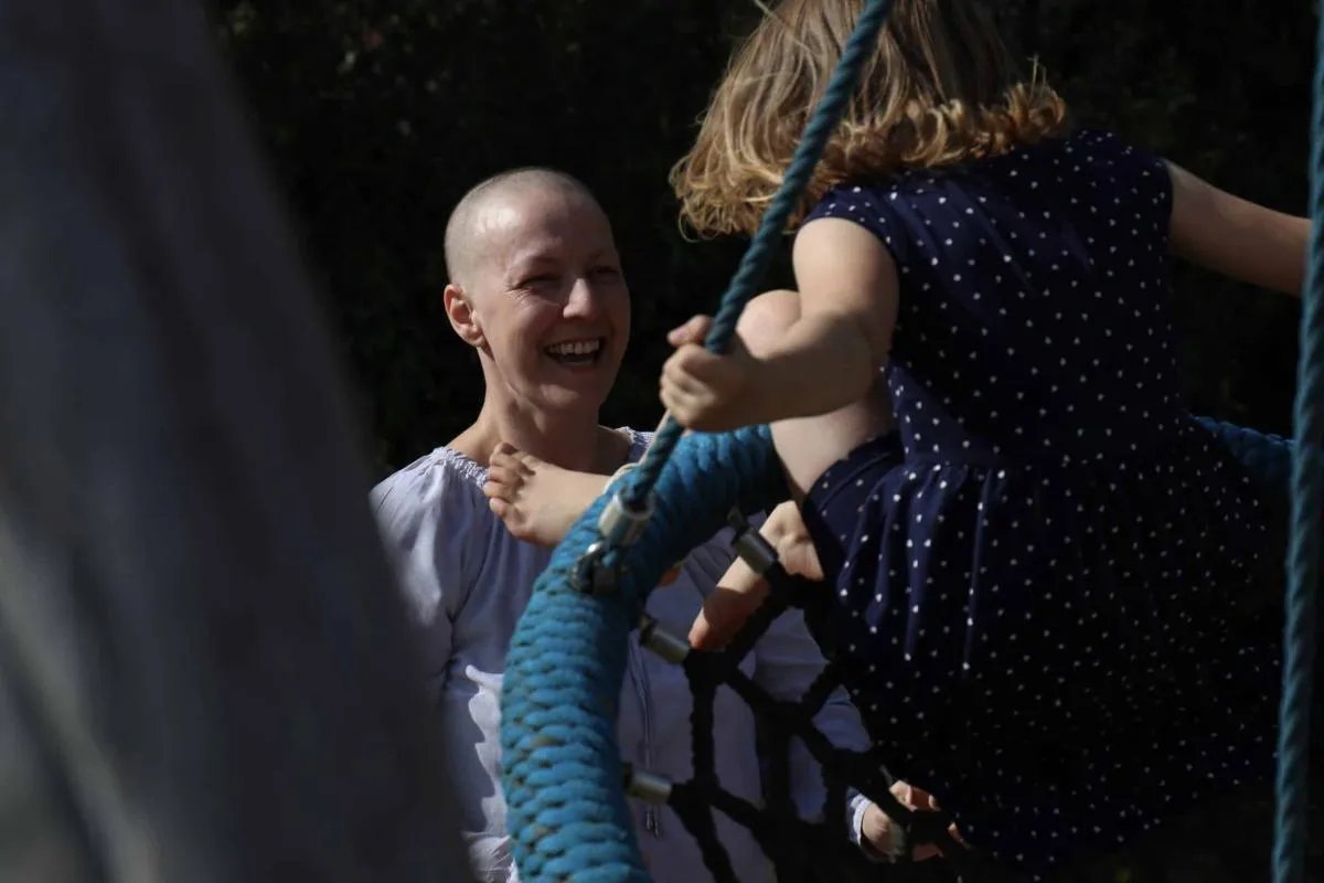 Frau mit kurzgeschorenem Haar lacht herzlich, während ein Kind im Pünktchenkleid auf einer Nestschaukel klettert.