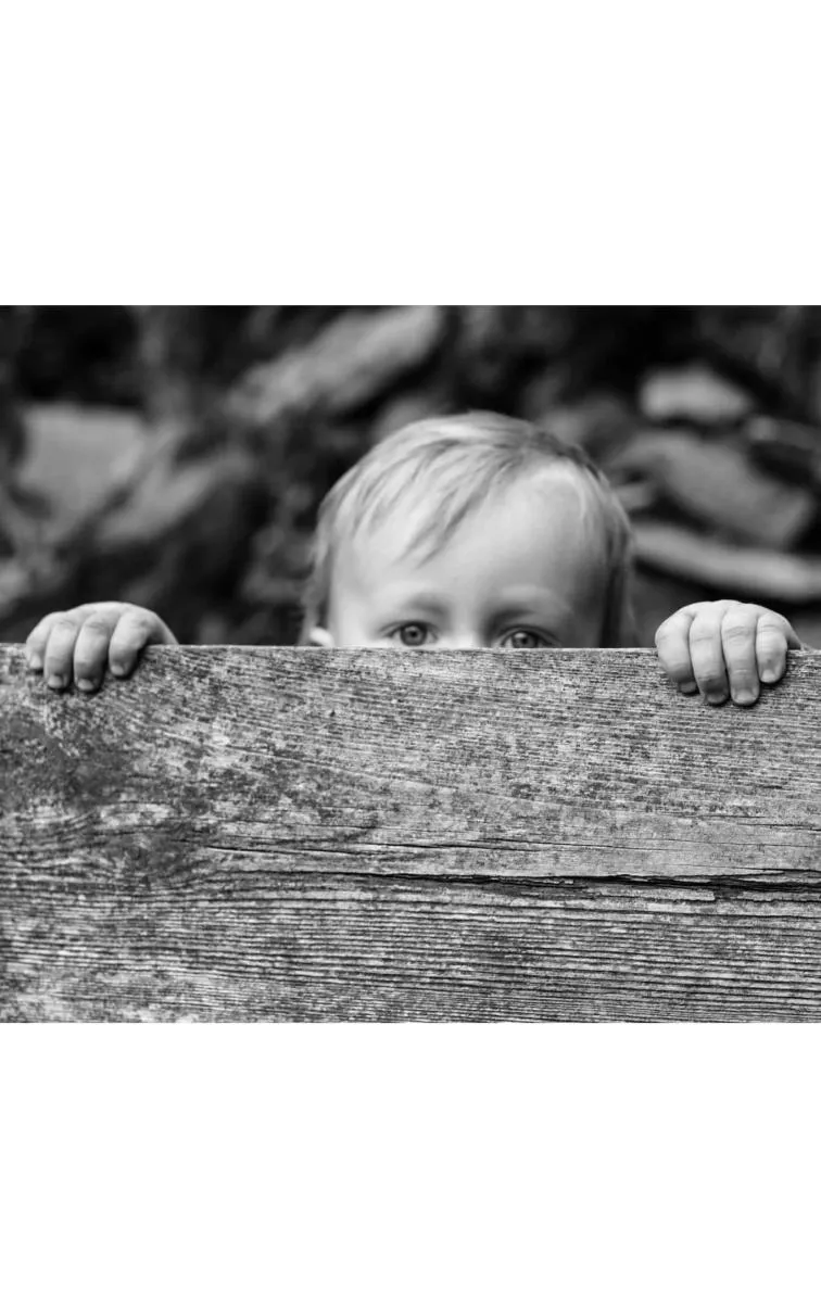 Schwarz-Weiß-Foto: Ein Kleinkind schaut neugierig über den Rand einer Holzplanke, nur Augen und Hände sichtbar.